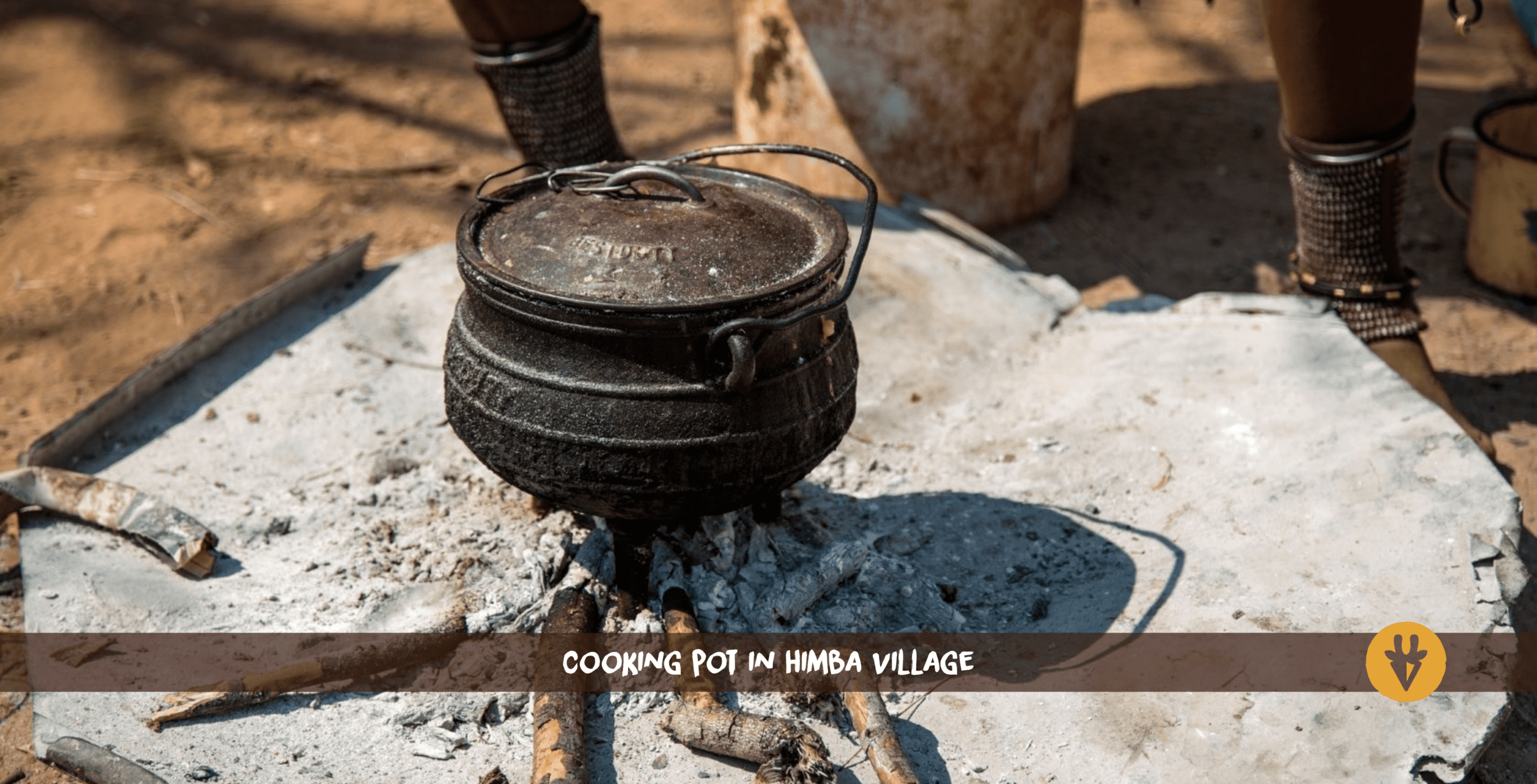 Un pot marron foncé avec couvercle et poignée posé sur de la cendre et des petites branches d'un ancien feu dans un village Himba.