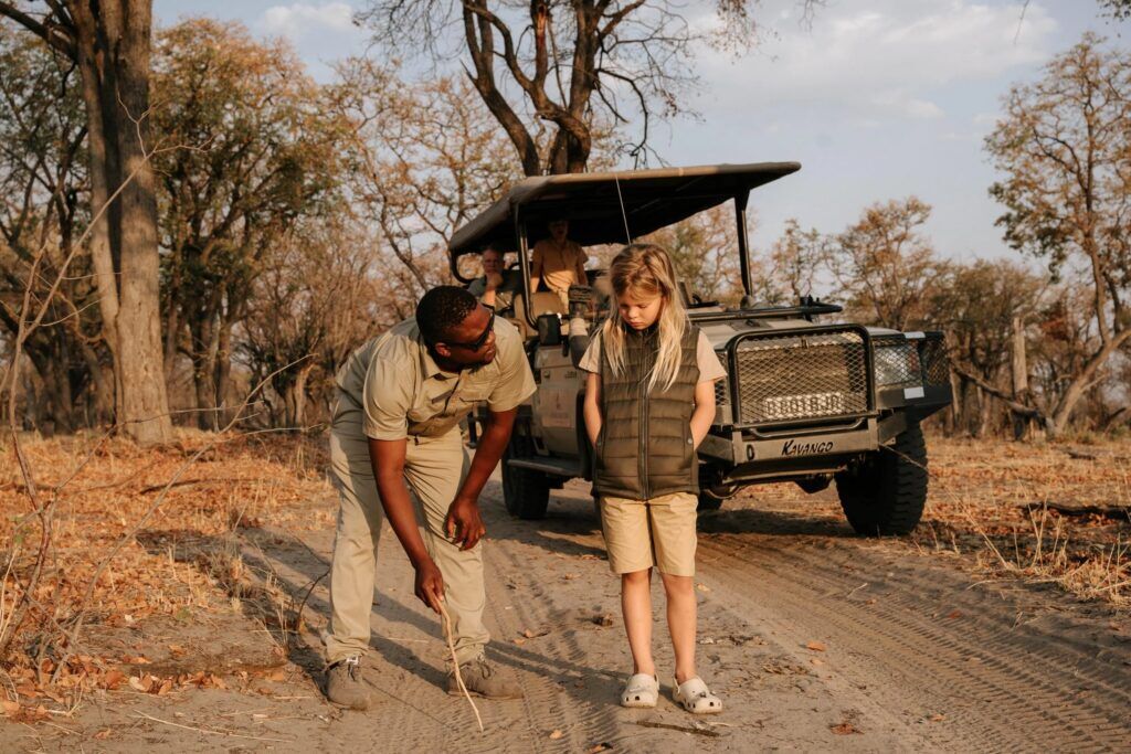 A man shows animal tracks in the dusty road to a young girl with a safari jeep in the background