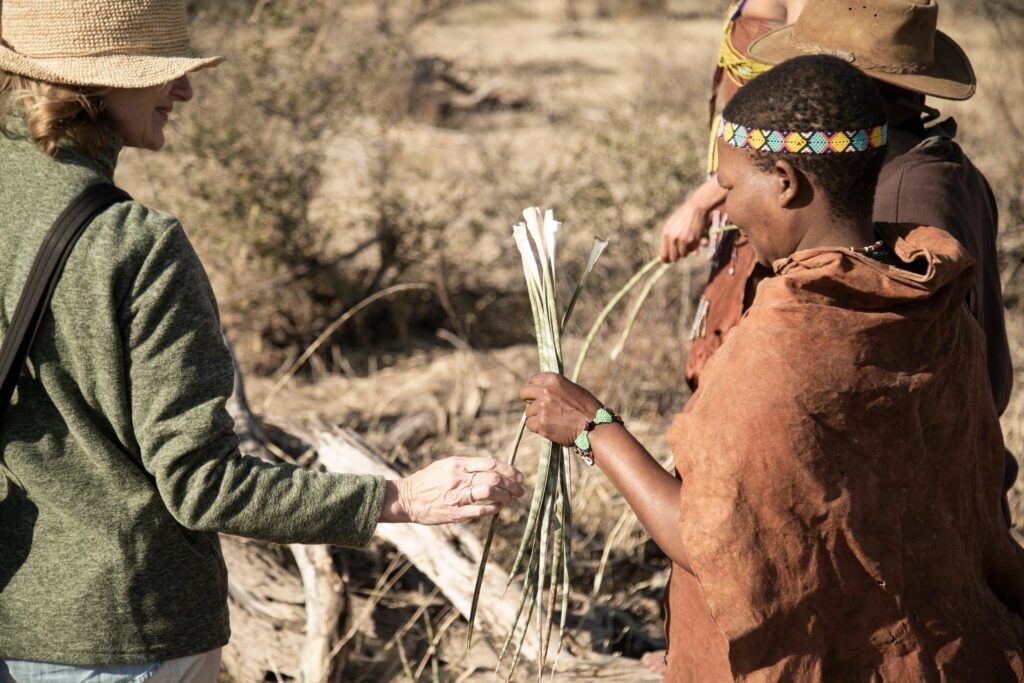 A woman with a hat and a San woman stand next to each other, the latter holding grass in her hands